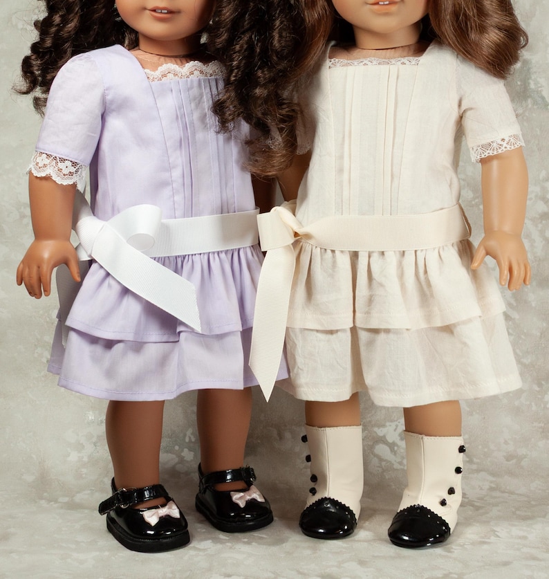 May include: Two dolls wearing matching dresses. One doll is in a light purple dress with a white ribbon belt and black shoes. The other doll is in an off-white dress with a cream ribbon belt and black and white boots.
