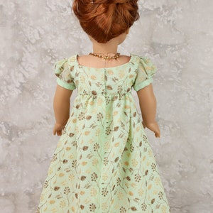 May include: A doll wearing a mint green dress with a gold leaf pattern. The dress has short sleeves and a gathered waist. The doll has red hair styled in a bun and wears a gold necklace. The dress is suitable for a doll.