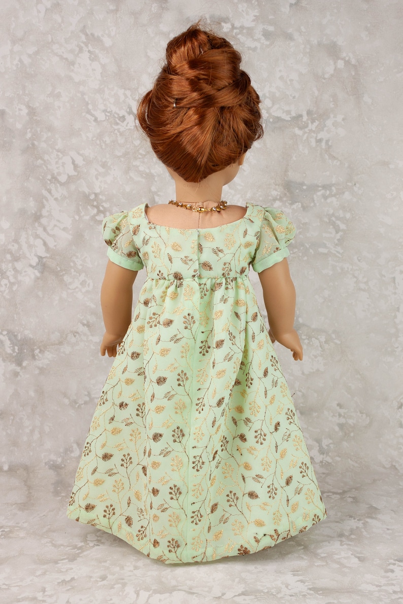 May include: A doll wearing a mint green dress with a gold leaf pattern. The dress has short sleeves and a gathered waist. The doll has red hair styled in a bun and wears a gold necklace. The dress is suitable for a doll.