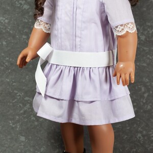 May include: A lavender doll dress with a white ribbon belt and lace trim. The dress features a tiered skirt and short sleeves. The doll is wearing black patent leather shoes with pink bows.