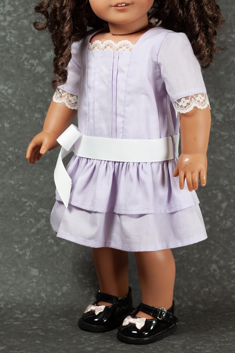 May include: A lavender doll dress with a white ribbon belt and lace trim. The dress features a tiered skirt and short sleeves. The doll is wearing black patent leather shoes with pink bows.