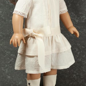 May include: A doll wearing a cream-colored dress with a layered skirt and a bow at the waist. The dress has short sleeves with lace trim. The doll is also wearing white boots with black accents and black buttons.