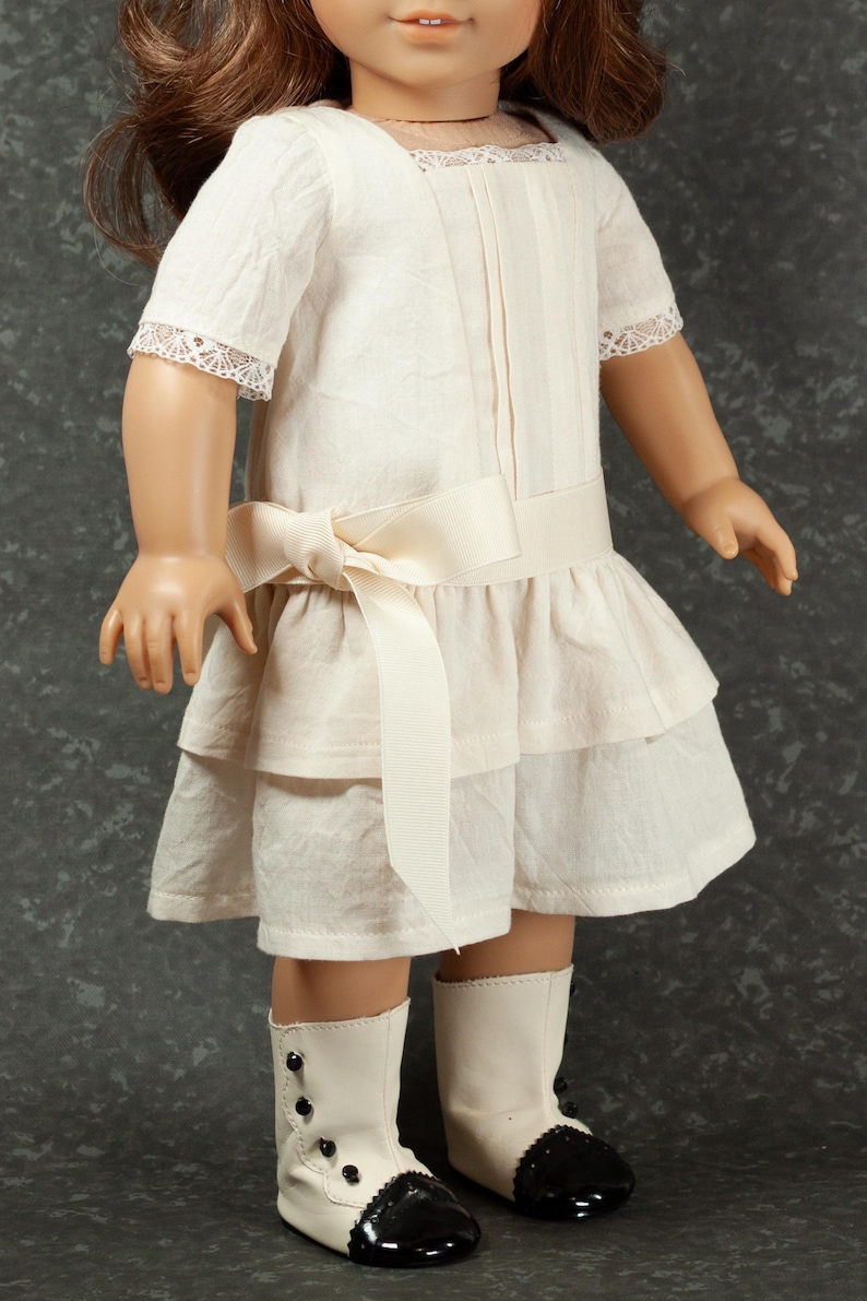 May include: A doll wearing a cream-colored dress with a layered skirt and a bow at the waist. The dress has short sleeves with lace trim. The doll is also wearing white boots with black accents and black buttons.
