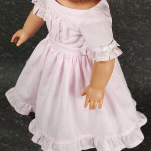 May include: A light pink doll dress featuring short ruffled sleeves with small white ribbon bows, a gathered waistline, and a ruffled hem. The dress has a round neckline and is worn by a doll with a light skin tone and a thin gold-colored necklace.