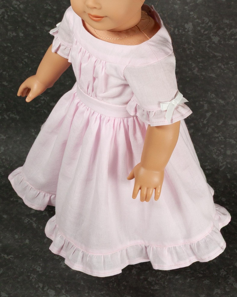 May include: A light pink doll dress featuring short ruffled sleeves with small white ribbon bows, a gathered waistline, and a ruffled hem. The dress has a round neckline and is worn by a doll with a light skin tone and a thin gold-colored necklace.