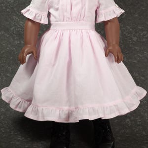 May include: A doll wearing a light pink dress with a boat neckline, short ruffled sleeves, and a gathered waist. The skirt features a wide hem with a ruffled trim. The doll is also wearing black lace-up shoes and has dark skin with brown eyes.