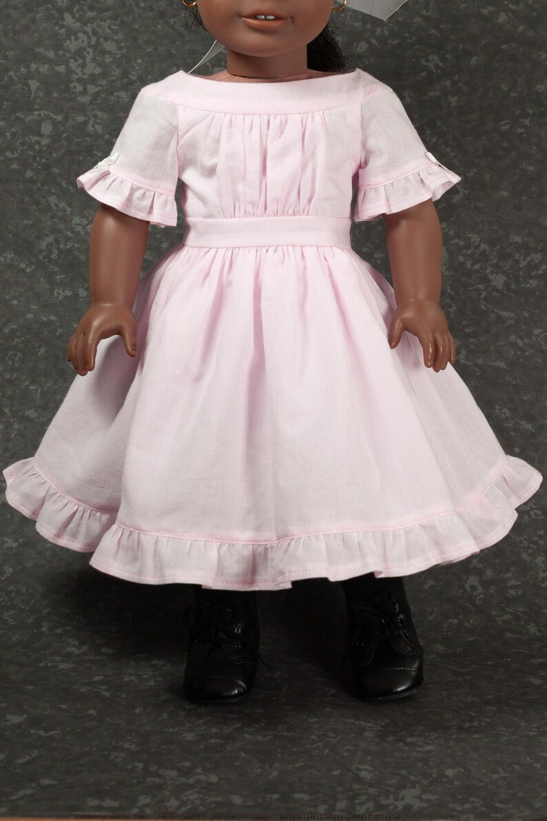 May include: A doll wearing a light pink dress with a boat neckline, short ruffled sleeves, and a gathered waist. The skirt features a wide hem with a ruffled trim. The doll is also wearing black lace-up shoes and has dark skin with brown eyes.