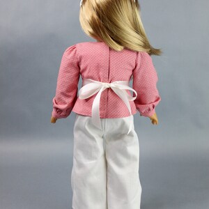 Puede incluir: Una mu&ntilde;eca con una blusa de estampado rosa y blanco con una faja blanca y pantalones blancos.