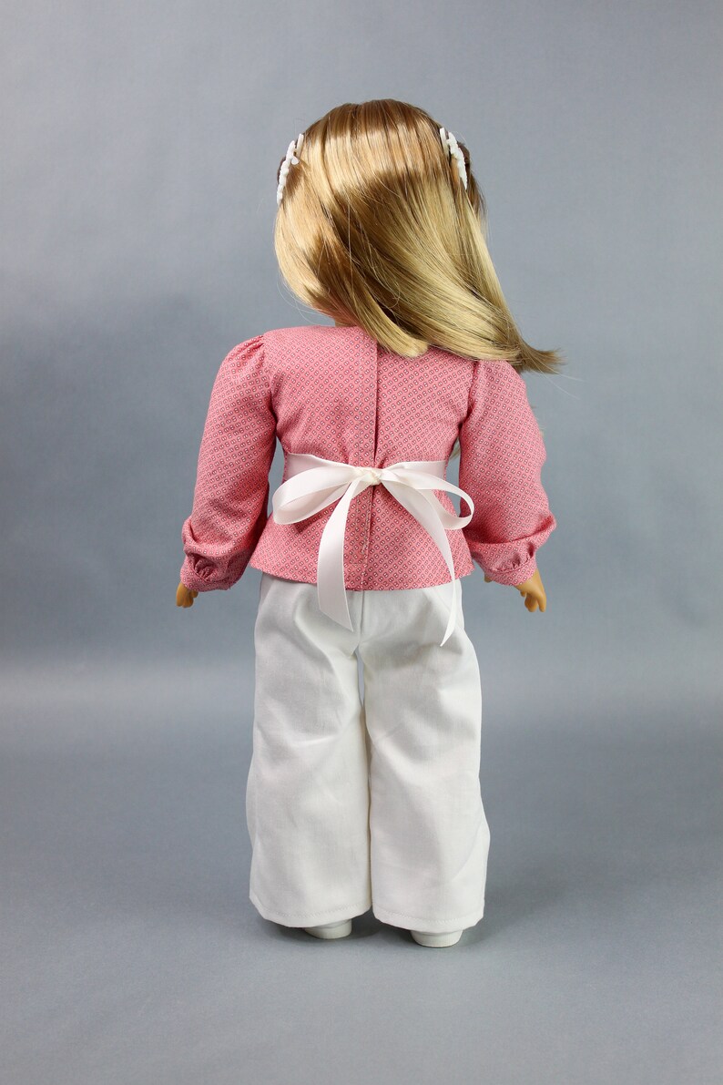 Puede incluir: Una mu&ntilde;eca con una blusa de estampado rosa y blanco con una faja blanca y pantalones blancos.