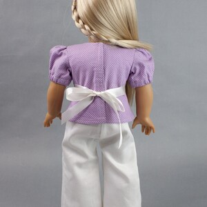 Puede incluir: Una mu&ntilde;eca con una blusa de lunares blancos y morados con una faja blanca y pantalones blancos.