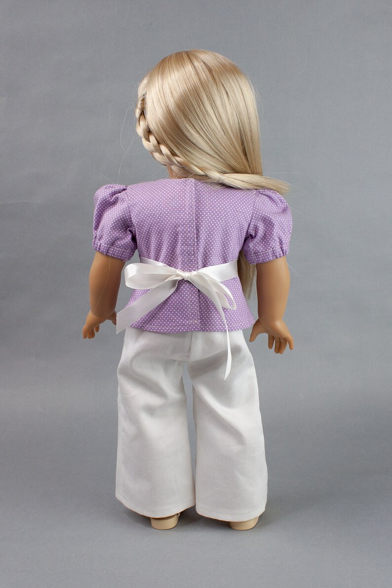 Puede incluir: Una mu&ntilde;eca con una blusa de lunares blancos y morados con una faja blanca y pantalones blancos.