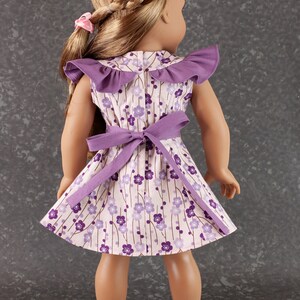 May include: A purple and white floral dress with a ruffled collar and a purple sash. The dress is worn by a doll with long brown hair.