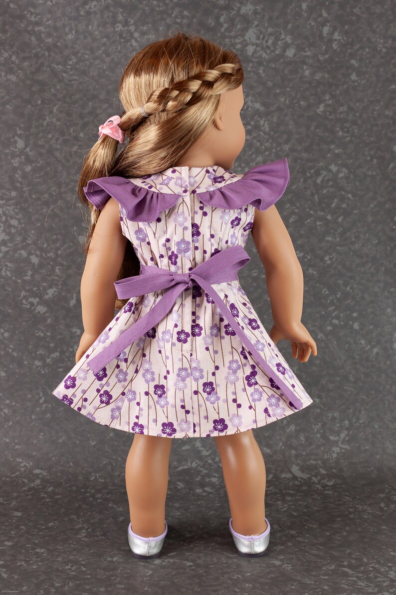 May include: A purple and white floral dress with a ruffled collar and a purple sash. The dress is worn by a doll with long brown hair.