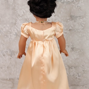 May include: A doll wearing a peach-colored satin dress with short, puffed sleeves and a square neckline. The doll has dark hair styled in an updo and is wearing a pearl necklace with a gold and green pendant. The dress has a fitted bodice and a flowing skirt.