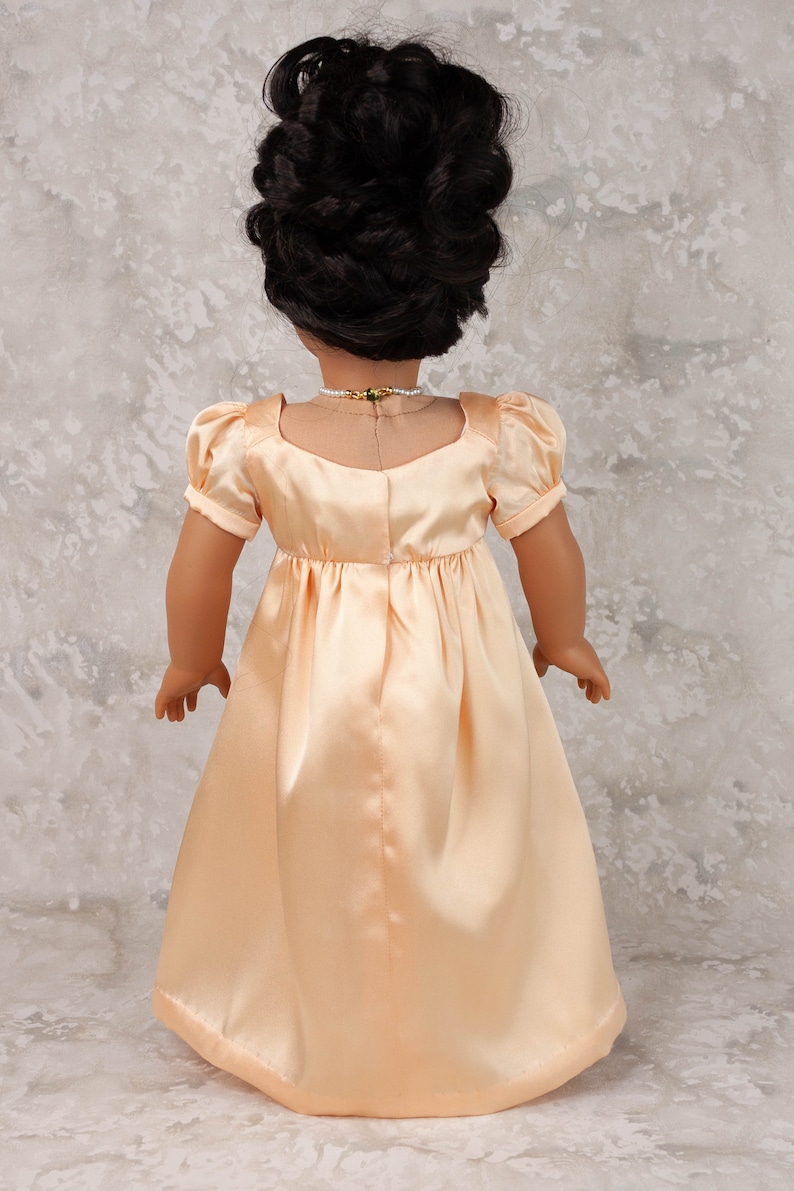 May include: A doll wearing a peach-colored satin dress with short, puffed sleeves and a square neckline. The doll has dark hair styled in an updo and is wearing a pearl necklace with a gold and green pendant. The dress has a fitted bodice and a flowing skirt.