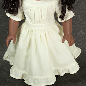 May include: A doll with dark curly hair wearing a cream-colored dress. The dress features a round neckline, short sleeves with ruffled edges, a fitted waistband, and a gathered skirt that ends in a wide ruffled hemline made of light fabric.