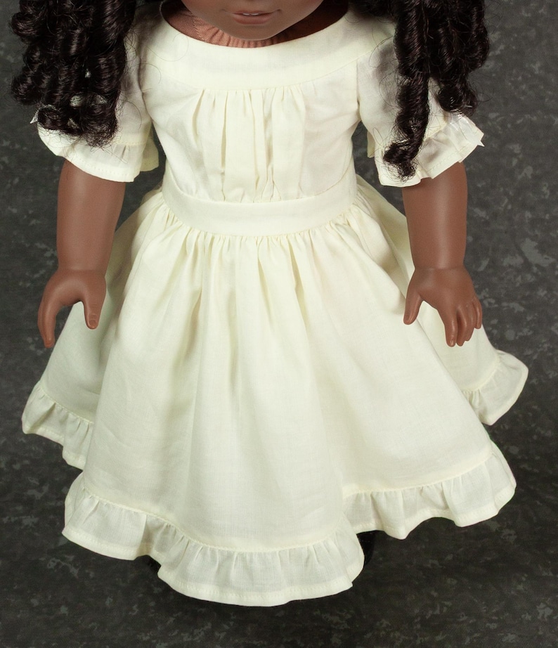 May include: A doll with dark curly hair wearing a cream-colored dress. The dress features a round neckline, short sleeves with ruffled edges, a fitted waistband, and a gathered skirt that ends in a wide ruffled hemline made of light fabric.