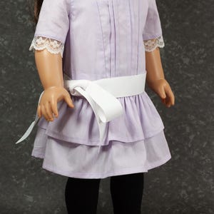 May include: A lavender doll dress with a white ribbon belt and lace trim. The dress features a square neckline and short sleeves with lace cuffs. The skirt has a layered design, and the doll is wearing black tights and black shoes.