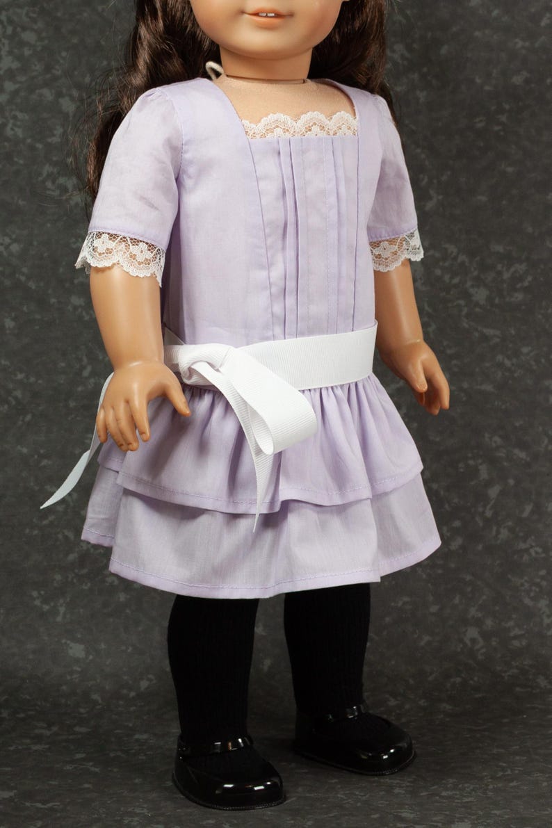May include: A lavender doll dress with a white ribbon belt and lace trim. The dress features a square neckline and short sleeves with lace cuffs. The skirt has a layered design, and the doll is wearing black tights and black shoes.
