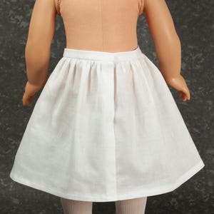May include: A doll wearing a sleeveless peach-colored top and a plain white knee-length skirt with a gathered waistband. The doll has light skin, reddish-brown hair, beige tights, and black shoes, viewed from the back against a dark gray background.