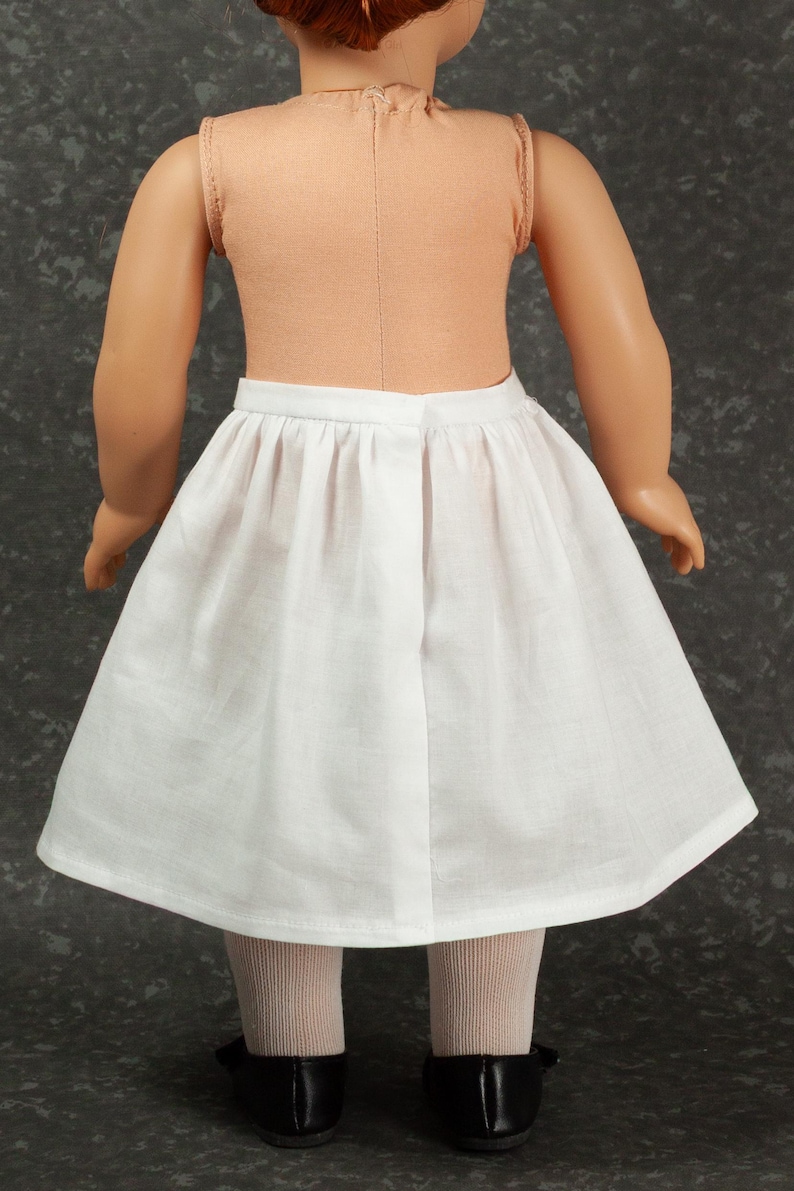 May include: A doll wearing a sleeveless peach-colored top and a plain white knee-length skirt with a gathered waistband. The doll has light skin, reddish-brown hair, beige tights, and black shoes, viewed from the back against a dark gray background.