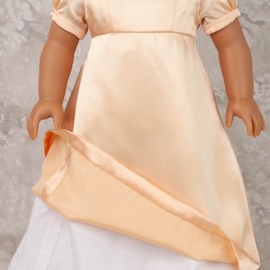 May include: A doll wearing a peach-colored satin dress with short, puffed sleeves and a white underskirt. The dress has a fitted bodice and a flowing skirt. A pearl necklace with a pendant adorns the doll's neck. The doll's shoes are dark purple.