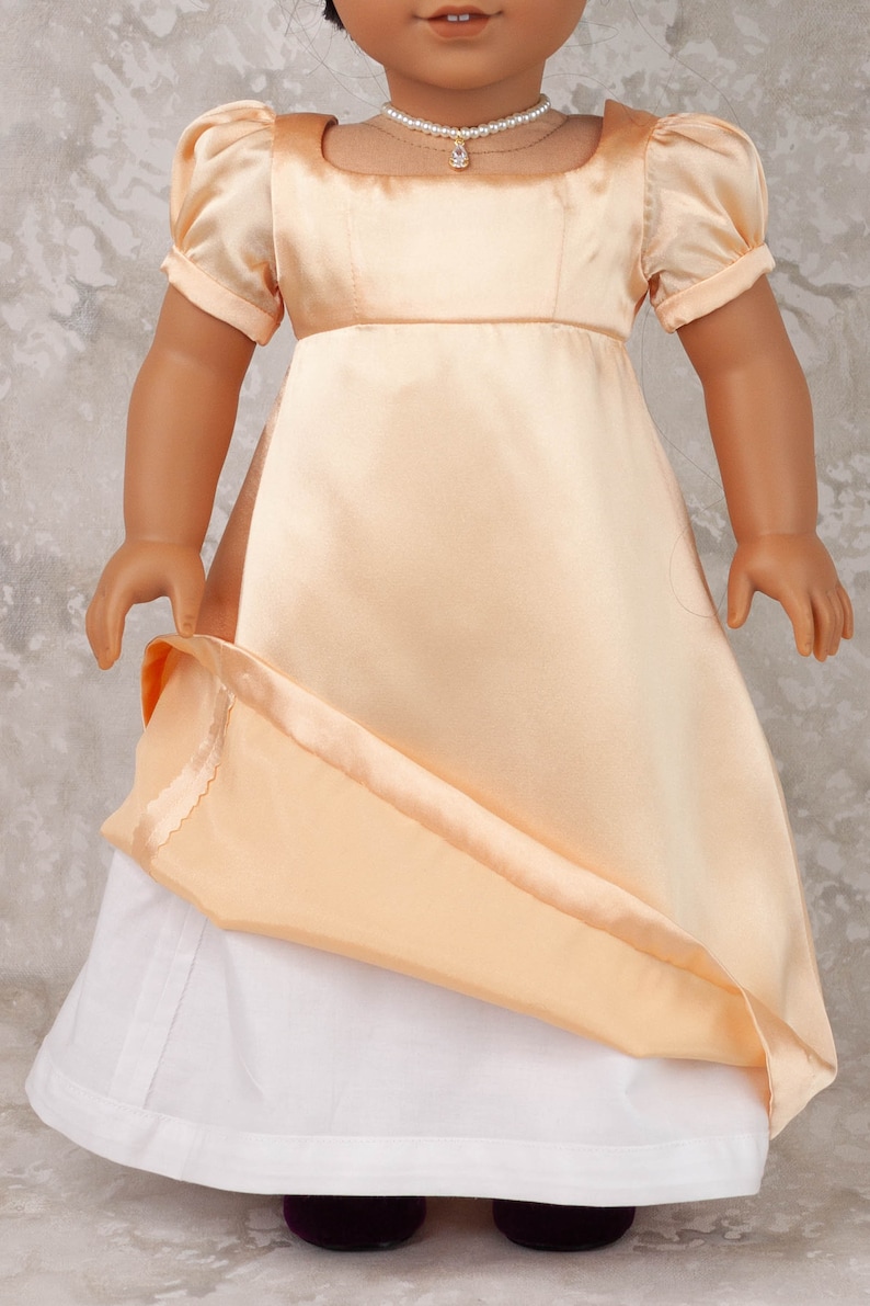 May include: A doll wearing a peach-colored satin dress with short, puffed sleeves and a white underskirt. The dress has a fitted bodice and a flowing skirt. A pearl necklace with a pendant adorns the doll's neck. The doll's shoes are dark purple.