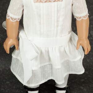 May include: A white doll dress with a square neckline, short sleeves, and a tiered skirt. The dress features lace trim and a white ribbon belt. The doll is wearing black patent leather shoes with pink bows.