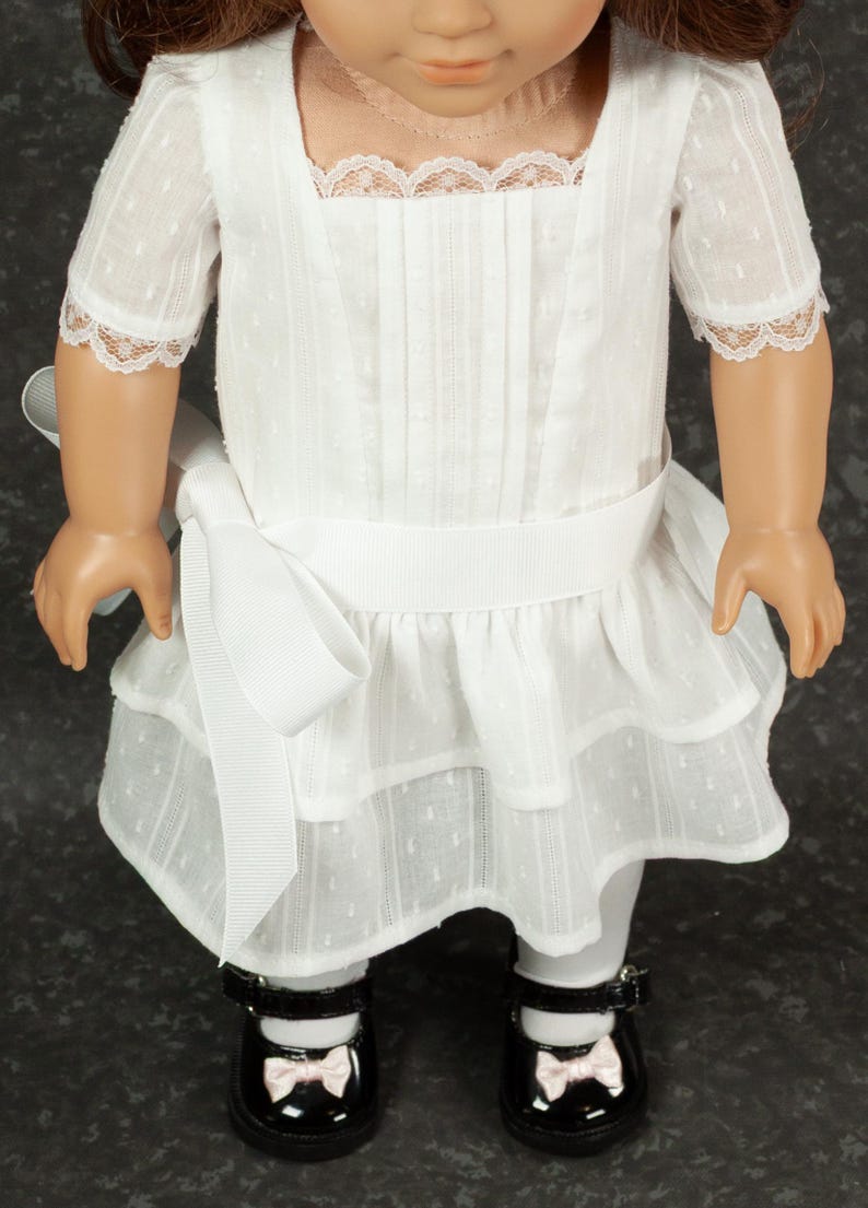 May include: A white doll dress with a square neckline, short sleeves, and a tiered skirt. The dress features lace trim and a white ribbon belt. The doll is wearing black patent leather shoes with pink bows.