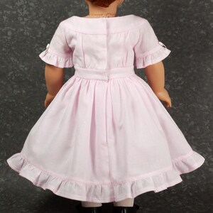 May include: A light pink dress worn by a doll, featuring short sleeves with ruffled edges, a fitted bodice with a back closure, a defined waistline, and a full skirt with a ruffled hem. The doll has auburn hair and wears black shoes.
