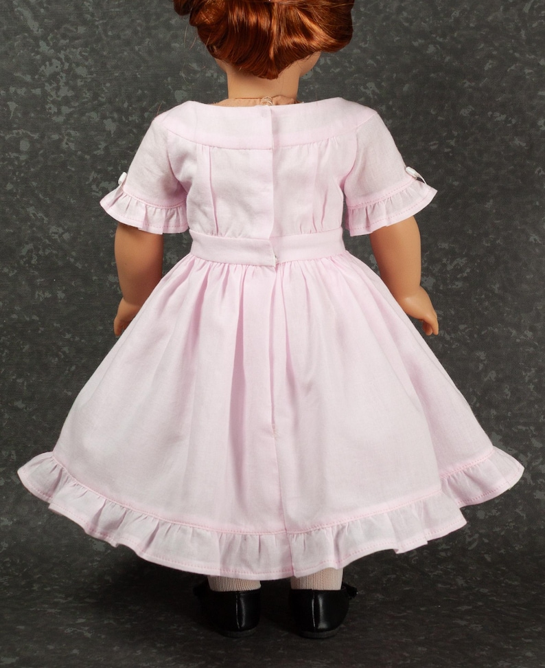 May include: A light pink dress worn by a doll, featuring short sleeves with ruffled edges, a fitted bodice with a back closure, a defined waistline, and a full skirt with a ruffled hem. The doll has auburn hair and wears black shoes.
