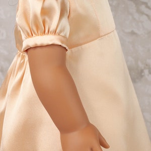 May include: A peach-colored satin doll dress with short, puffed sleeves. The dress features a fitted bodice and a flowing skirt. A pearl necklace with a gold pendant is visible around the doll's neck. The doll's arm and hand are also visible.