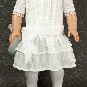 May include: A white doll dress with a square neckline, short sleeves with lace trim, and a tiered skirt. The dress has a white ribbon belt and a large bow on the side. The doll is wearing white tights and black patent leather shoes with pink bows.