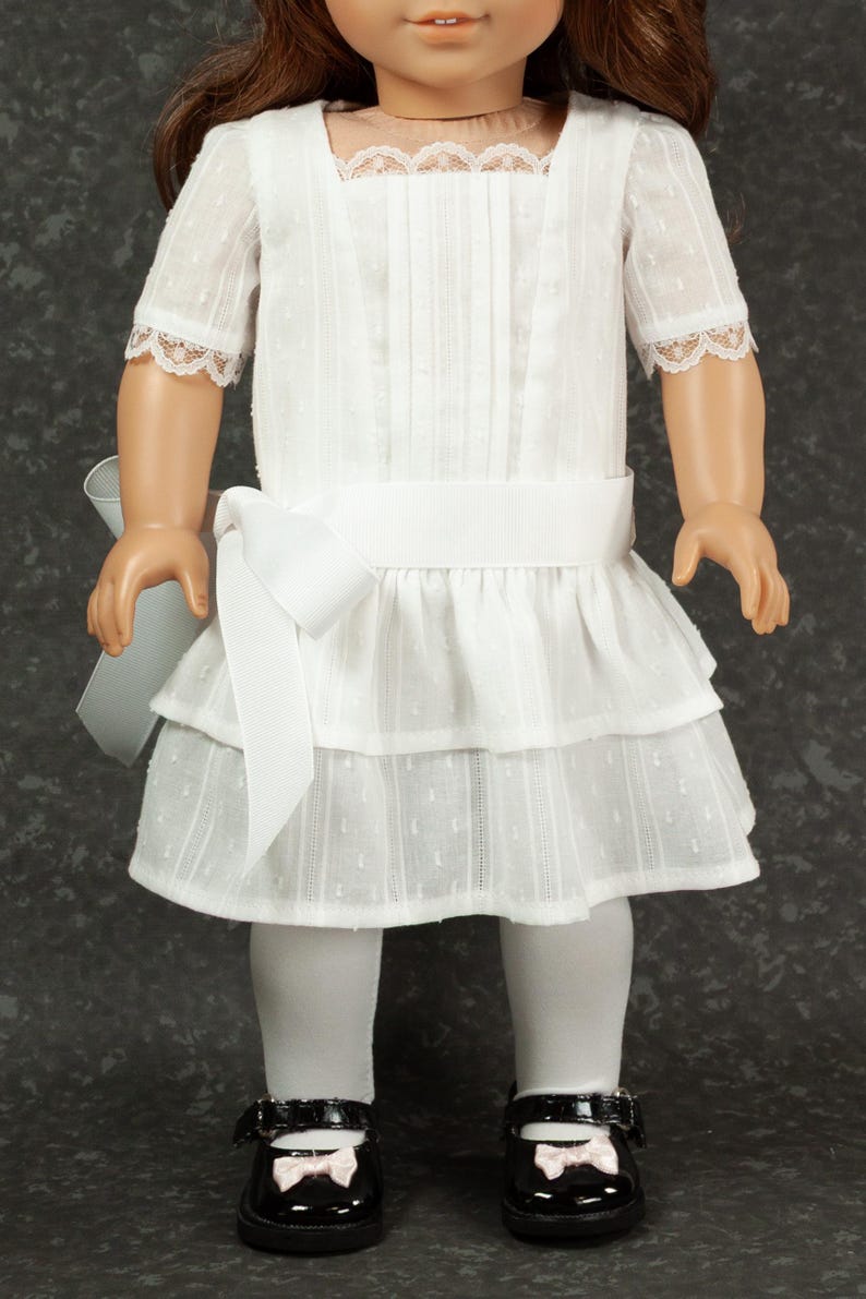 May include: A white doll dress with a square neckline, short sleeves with lace trim, and a tiered skirt. The dress has a white ribbon belt and a large bow on the side. The doll is wearing white tights and black patent leather shoes with pink bows.