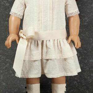 May include: A cream-colored doll dress with a square neckline, short sleeves with lace trim, and a tiered skirt. A cream-colored ribbon belt and bow accent the waist. The doll is wearing white boots with black accents.