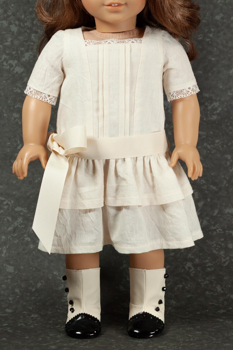 May include: A cream-colored doll dress with a square neckline, short sleeves with lace trim, and a tiered skirt. A cream-colored ribbon belt and bow accent the waist. The doll is wearing white boots with black accents.