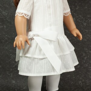 May include: A doll wearing a white dress with a tiered skirt, short sleeves, and a bow at the waist. The dress has a textured pattern and lace trim. The doll is also wearing white tights and black patent leather shoes with pink bows.
