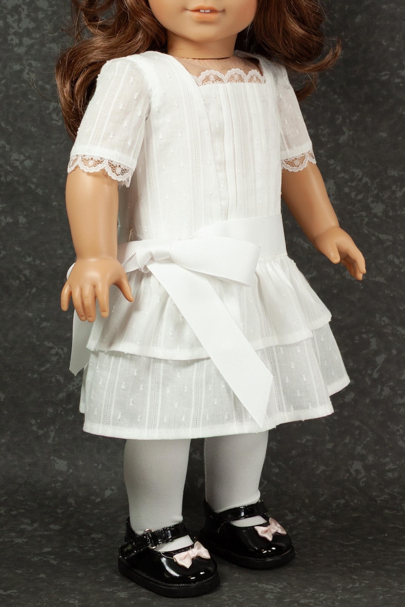 May include: A doll wearing a white dress with a tiered skirt, short sleeves, and a bow at the waist. The dress has a textured pattern and lace trim. The doll is also wearing white tights and black patent leather shoes with pink bows.