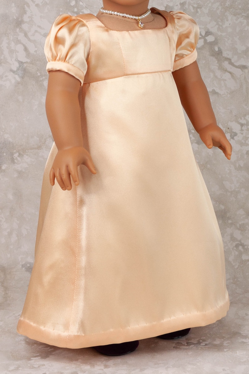 May include: A peach-colored satin doll dress with short, puffed sleeves and a square neckline. The dress features a fitted bodice and a flowing skirt. A pearl necklace with a pendant is visible.