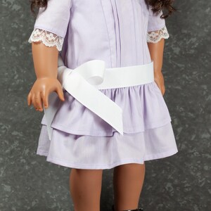 May include: A lavender doll dress with a white ribbon belt and lace trim on the sleeves and neckline. The dress has a tiered skirt and is paired with black patent leather shoes with white bows.