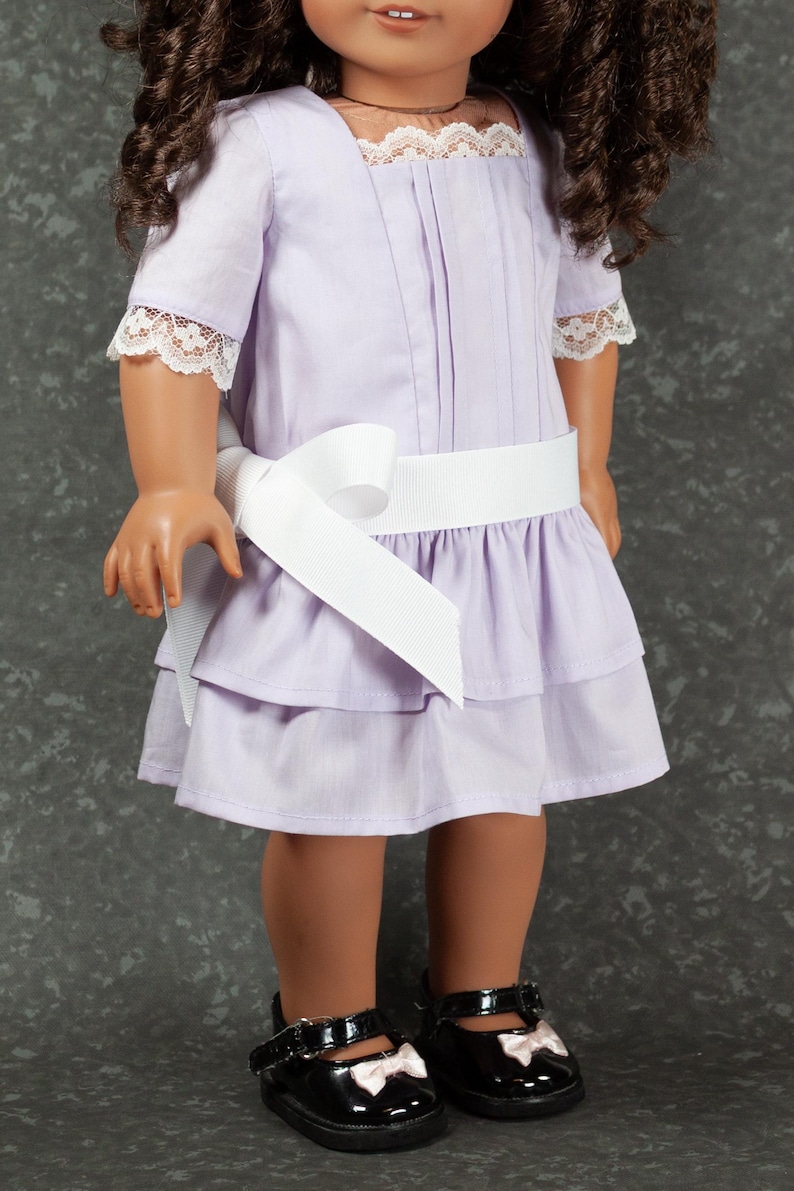 May include: A lavender doll dress with a white ribbon belt and lace trim on the sleeves and neckline. The dress has a tiered skirt and is paired with black patent leather shoes with white bows.