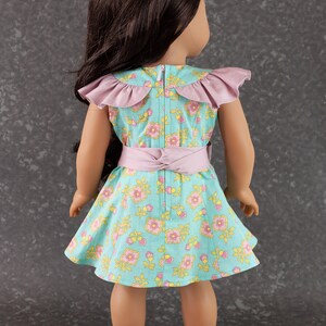 May include: A doll wearing a light blue and pink floral dress with a pink ruffled collar and a pink sash. The doll has long brown hair and is wearing sandals.