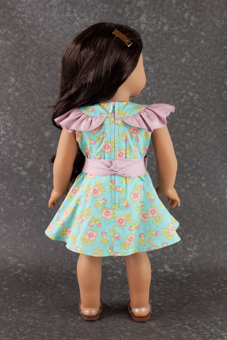 May include: A doll wearing a light blue and pink floral dress with a pink ruffled collar and a pink sash. The doll has long brown hair and is wearing sandals.