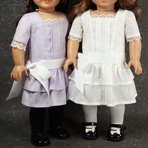 May include: Two dolls wearing matching dresses. One doll is in a lavender dress with black tights and black shoes. The other doll is in a white dress with white tights and black shoes. Both dresses have a white bow and lace trim.
