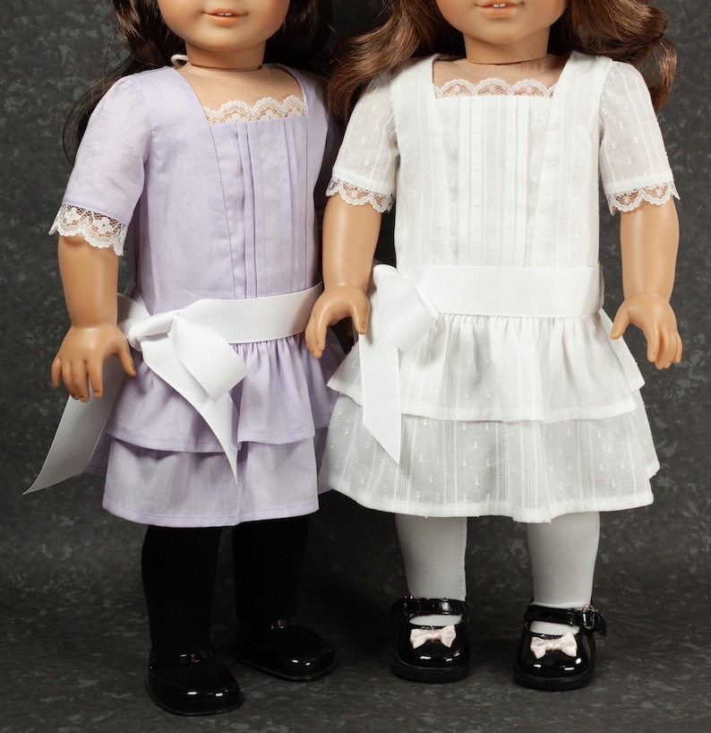 May include: Two dolls wearing matching dresses. One doll is in a lavender dress with black tights and black shoes. The other doll is in a white dress with white tights and black shoes. Both dresses have a white bow and lace trim.