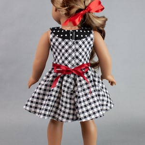 May include: A black and white gingham dress with red ribbon accents. The dress has a white polka dot collar and a fitted bodice. The doll is wearing black shoes.