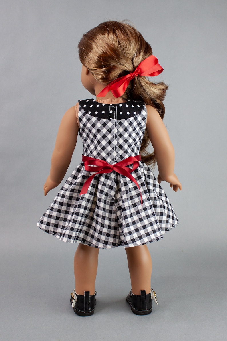 May include: A black and white gingham dress with red ribbon accents. The dress has a white polka dot collar and a fitted bodice. The doll is wearing black shoes.