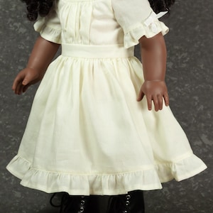 May include: A doll with dark curly hair wearing a cream-colored dress featuring short sleeves with small white bows, a gathered bodice, a defined waistline, and a ruffled hemline, paired with black lace-up boots that have silver-toned eyelets.