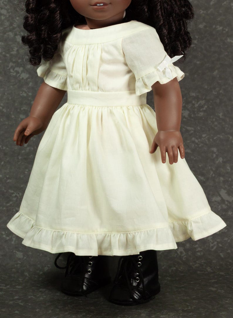 May include: A doll with dark curly hair wearing a cream-colored dress featuring short sleeves with small white bows, a gathered bodice, a defined waistline, and a ruffled hemline, paired with black lace-up boots that have silver-toned eyelets.