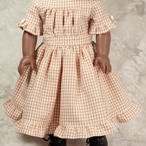 May include: A doll wearing a tan and white gingham dress with a gathered bodice, short ruffled sleeves, and a ruffled hemline. The dress features a fitted waistband and is paired with black lace-up shoes. The doll has dark skin and dark hair.